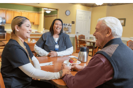 Nurses chatting with a resident at the dining table.